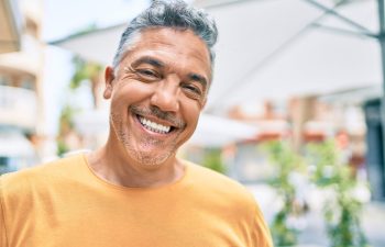 A smiling man in a yellow shirt stands outdoors under umbrellas, with blurred greenery and buildings in the background.